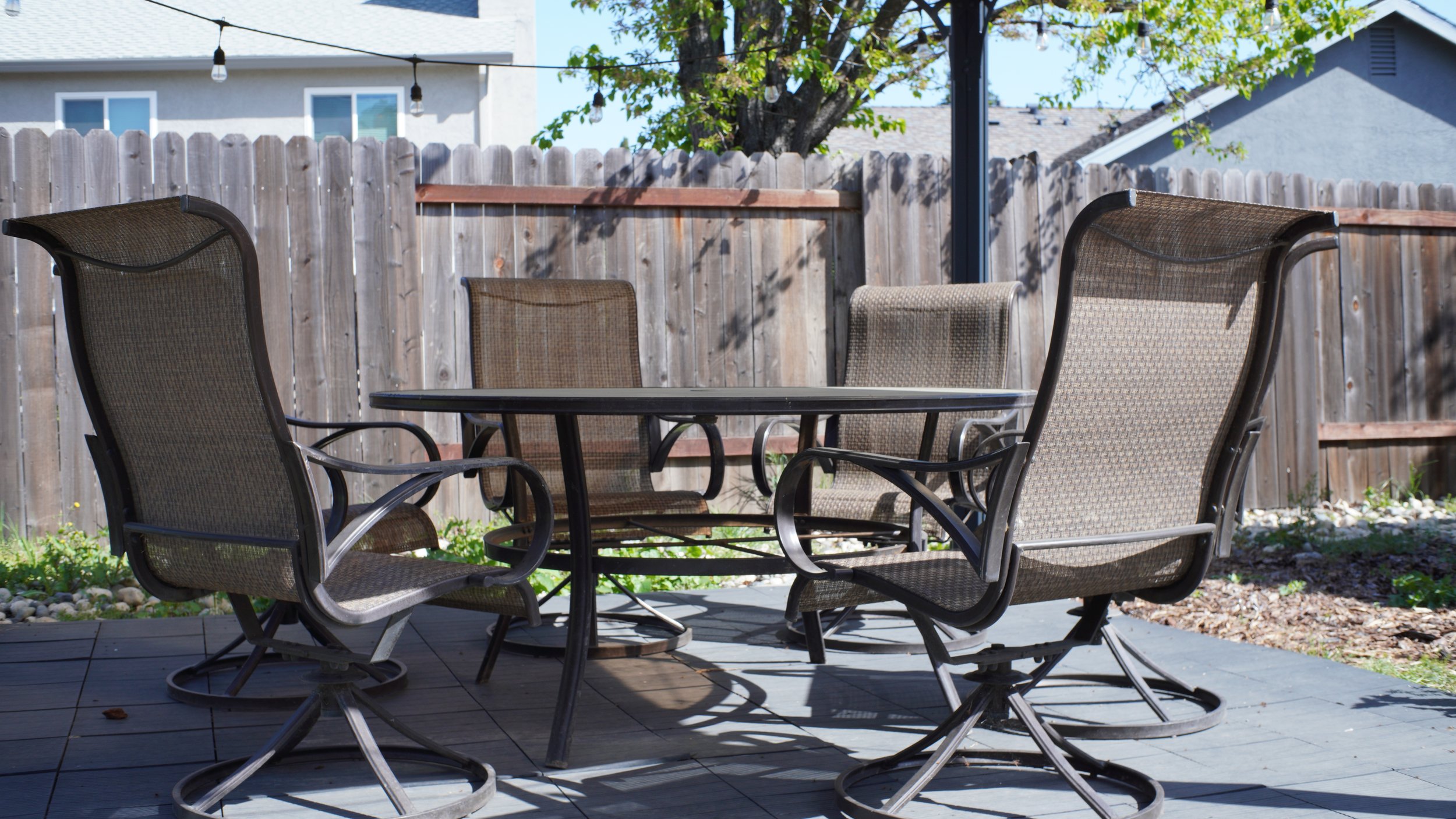 The backyard patio at the Elk Grove house: a shaded table with four mesh patio chairs against a wood fence, string lights overhead